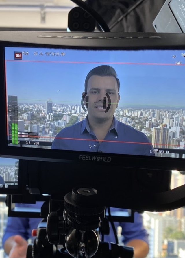 Monitor da câmera mostrando Daniel falando, com a cidade ao fundo